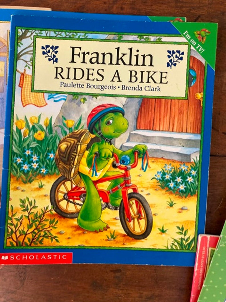 K&ouml;nnte beinhalten: Ein Kinderbuchcover mit einer gr&uuml;nen Schildkr&ouml;te, die einen Helm tr&auml;gt und ein rotes Fahrrad f&auml;hrt. Der Buchtitel lautet "Franklin Rides a Bike" von Paulette Bourgeois und Brenda Clark. Das Cover zeigt au&szlig;erdem einen blauen Himmel, gr&uuml;nes Gras und gelbe Blumen.
