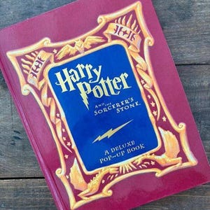 May include: A deluxe pop-up book titled "Harry Potter and the Sorcerer's Stone." The book has a burgundy cover with gold and blue accents. The title is in a stylized font, and the cover features decorative elements.