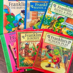 K&ouml;nnte beinhalten: Ein Stapel von sechs bunten Kinderb&uuml;chern mit einer Schildkr&ouml;tenfigur namens Franklin. Die B&uuml;cher tragen die Titel "Franklin und die Babysitterin", "Franklin m&ouml;chte ein Haustier", "Franklin f&auml;hrt Fahrrad", "Franklin und das Schulst&uuml;ck", "Franklin ist der Boss" und "Franklin und der Zauber-Stift".
