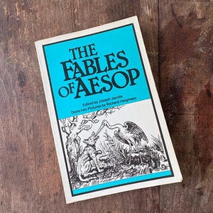 May include: A hardcover book titled The Fables of Aesop, edited by Joseph Jacobs and illustrated by Richard Heighway. The cover features a bright turquoise top section with black text and a white bottom section with a black and white illustration.