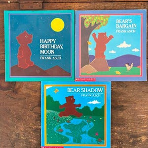 May include: Three children's books by Frank Asch are displayed. The books feature illustrations of a bear in various scenes. The titles include "Happy Birthday, Moon," "Bear's Bargain," and "Bear Shadow." Each book has a colorful cover with a distinct design.