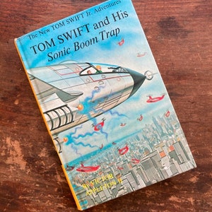 May include: A yellow and white book cover with a blue and white illustration of a jet plane flying over a city. The book title is "The New Tom Swift Jr. Adventures Tom Swift and His Sonic Boom Trap" by Victor Appleton II.