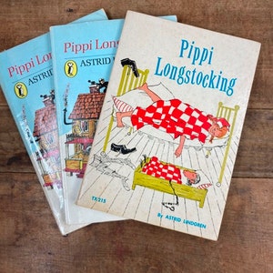 May include: Three vintage paperback books with colorful covers featuring illustrations of Pippi Longstocking, a fictional character created by Astrid Lindgren. The books are titled "Pippi Longstocking" and feature a red and white checkered blanket, a bed, and a girl with pigtails.