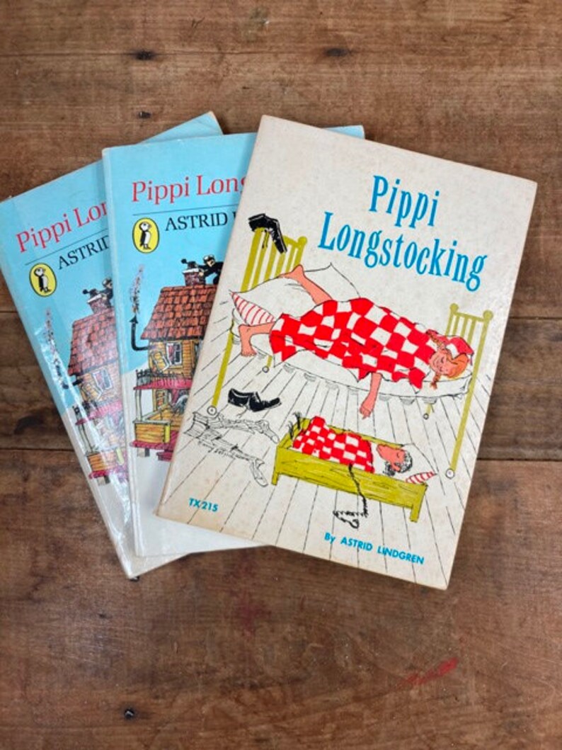 May include: Three vintage paperback books with colorful covers featuring illustrations of Pippi Longstocking, a fictional character created by Astrid Lindgren. The books are titled "Pippi Longstocking" and feature a red and white checkered blanket, a bed, and a girl with pigtails.
