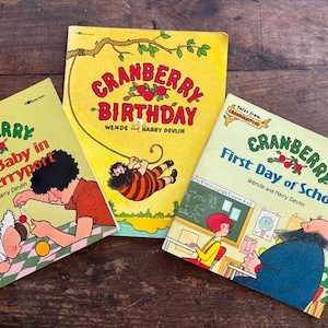 May include: Three vintage children's books with colorful illustrations. The titles are "Cranberry Birthday", "Cranberry First Day of School", and "Cranberry A New Baby in Cranberryport". The books have yellow and light green covers with red text and illustrations.