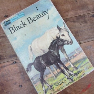 May include: A vintage book cover featuring a black and white horse with a foal. The book title is "Black Beauty" by Anna Sewell. The cover is illustrated by E. Raymond Kinstler.