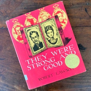 May include: A red hardcover book with a gold sticker on the front cover. The book title is "They Were Strong and Good" by Robert Lawson. The cover features illustrations of people in frames.