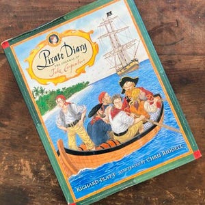 May include: A green and red book titled "Pirate Diary: The Journal of Jake Carpenter" by Richard Platt, illustrated by Chris Riddell. The cover features a pirate ship sailing on the ocean with a group of pirates in a rowboat.