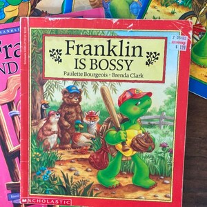 K&ouml;nnte beinhalten: Ein rotes Kinderbuch mit dem Titel "Franklin ist ein Tyrann" von Paulette Bourgeois und Brenda Clark. Das Cover zeigt eine gr&uuml;ne Schildkr&ouml;te, die eine rote Baseballkappe tr&auml;gt und einen Baseballschl&auml;ger und einen Handschuh h&auml;lt. Die Schildkr&ouml;te geht auf einem Weg mit anderen Tieren, darunter ein Fuchs, ein B&auml;r und ein Eichh&ouml;rnchen.