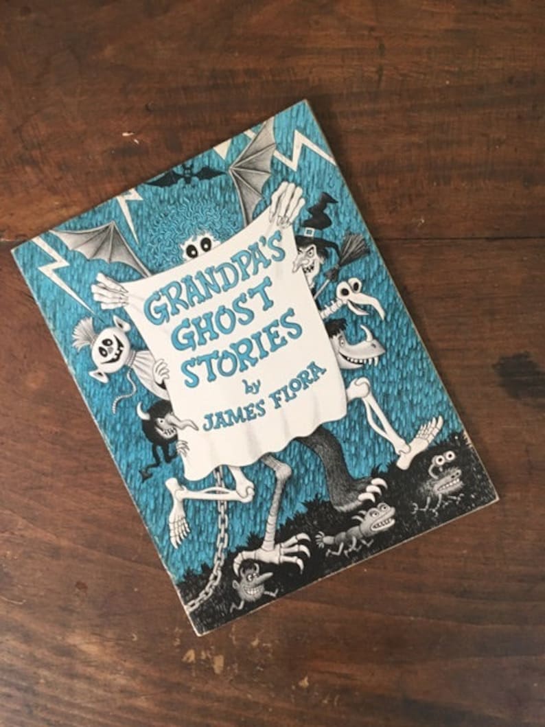 Pode incluir: Uma capa de livro azul com o t&iacute;tulo "Grandpa's Ghost Stories" de James Flora. A capa apresenta um esqueleto segurando um len&ccedil;ol com o t&iacute;tulo e uma variedade de personagens assustadores.