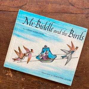 May include: A vintage children's book titled "Mr. Biddle and the Birds" by Lonzo Anderson, illustrated by Adrienne Adams. The cover features a blue sky background with illustrations of birds and a whimsical scene. The book is set on a wooden surface.