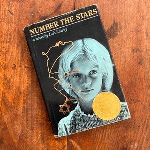 May include: A black paperback book with the title "NUMBER THE STARS" and the subtitle "a novel by Lois Lowry". The cover features a black and white photo of a young girl with blonde hair wearing a necklace with a Star of David pendant. A gold medal is visible on the cover.