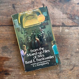May include: A hardcover book titled "From the Mixed-up Files of Mrs. Basil E. Frankweiler" by E.L. Konigsburg. The cover features an illustration of a museum scene with figures and text. The book is lying on a wooden surface.
