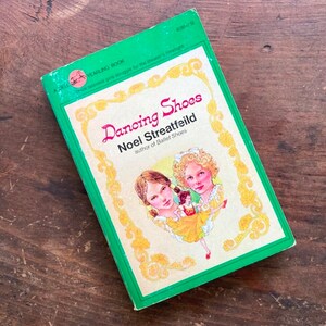 May include: A paperback book titled Dancing Shoes by Noel Streatfeild, published by Dell Yearling. The cover features a green border, a cream-colored center, and an illustration of two young people with a small figure in a yellow dress in the middle.