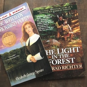 May include: Two paperback books, one titled "The Witch of Blackbird Pond" by Elizabeth George Speare and the other titled "The Light in the Forest" by Conrad Richter. Both books are on a wooden surface.