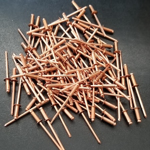 May include: A large pile of copper-colored rivets. The rivets are about 1 inch long and have a round head. They are arranged in a random pattern.