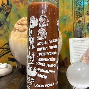 May include: A tall, clear glass candle filled with reddish-brown wax. The candle features white text and illustrations, including the words "DOBLE FUERZA" and images of Native American headdresses. The candle is on a wooden surface.