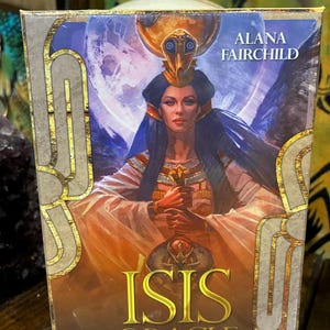 Könnte beinhalten: Die Box für das "Isis Oracle" Kartendeck von Alana Fairchild. Die Box zeigt ein Porträt einer Frau mit goldenem Kopfschmuck und Schwert, vor dem Hintergrund eines Vollmonds und blauer Flügel. Der Text "Isis Oracle" ist deutlich sichtbar.
