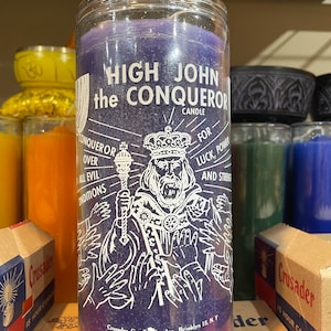 May include: A tall, cylindrical purple candle with the text "HIGH JOHN the CONQUEROR" in white. The candle features a detailed illustration of a crowned figure holding a scepter, surrounded by hands. The text also includes "FOR LUCK, POWER, AND STRENGTH".