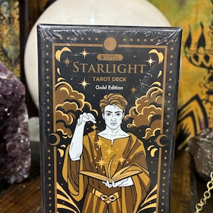 Puede incluir: Una caja negra y dorada "Wyspell Starlight Tarot Deck Gold Edition". La caja presenta una ilustración de una persona con una túnica dorada sosteniendo un libro, con estrellas y nubes. La caja está sellada en plástico transparente.