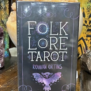 May include: A black tarot card box with the words "FOLKLORE TAROT" in white, and "ROWAN ORTINS" in white. The box features a purple owl illustration and decorative swirls. The background includes a crystal and wooden surface.