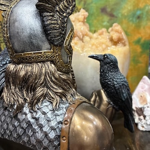 May include: A detailed sculpture of a warrior with a silver helmet, gold accents, and a bronze-colored chest. A black raven sits nearby. The sculpture has a textured, metallic appearance, suggesting a historical or fantasy theme.