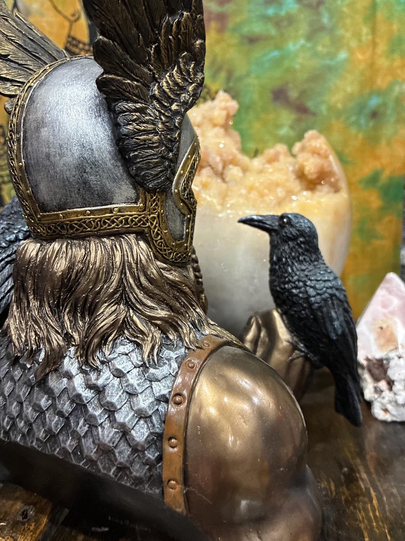 May include: A detailed sculpture of a warrior with a silver helmet, gold accents, and a bronze-colored chest. A black raven sits nearby. The sculpture has a textured, metallic appearance, suggesting a historical or fantasy theme.