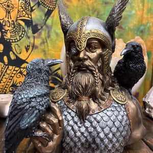 May include: A detailed bust of a Viking figure with a silver and gold helmet, a long beard, and a single eye. Two black ravens perch on the figure's shoulders and hand. The bust has a metallic finish and is set against a colorful backdrop.