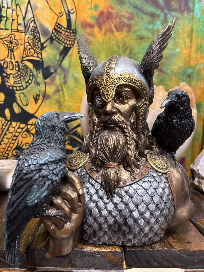 May include: A detailed bust of a Viking figure with a silver and gold helmet, a long beard, and a single eye. Two black ravens perch on the figure's shoulders and hand. The bust has a metallic finish and is set against a colorful backdrop.