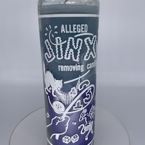 May include: A cylindrical glass candle with a dark blue label. The label reads "ALLEGED JINX removing candle" in white text, with illustrations of a cat, dice, and the number 13. The candle has a dark blue wax.