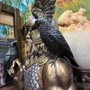 May include: A detailed bust of a Norse god with a raven perched on the shoulder. The statue features a bronze and silver color scheme, with intricate detailing on the helmet, beard, and raven's feathers. The bust is a decorative item.
