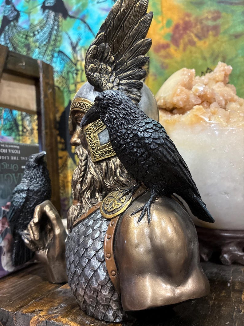 May include: A detailed bust of a Norse god with a raven perched on the shoulder. The statue features a bronze and silver color scheme, with intricate detailing on the helmet, beard, and raven's feathers. The bust is a decorative item.
