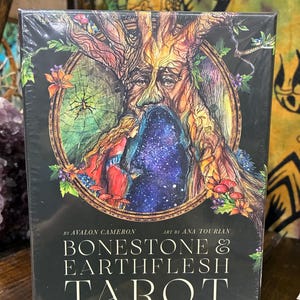 May include: A tarot card deck in a black box with the title "Bonestone & Earthflesh Tarot: The Reawakening." The box features a colorful illustration of a tree with a face, a woman, and a starry background.