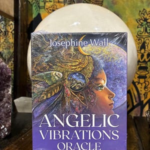 May include: A boxed oracle deck titled "Angelic Vibrations Oracle" by Josephine Wall. The box is purple with an illustration of a woman's profile with a celestial background. The deck includes a 50-card deck and guidebook.
