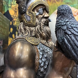 May include: A bronze-toned bust of a Viking warrior with a detailed helmet and beard. A raven sits on the warrior's shoulder. The bust has intricate gold-colored accents and a weathered appearance, suggesting a historical or fantasy theme.