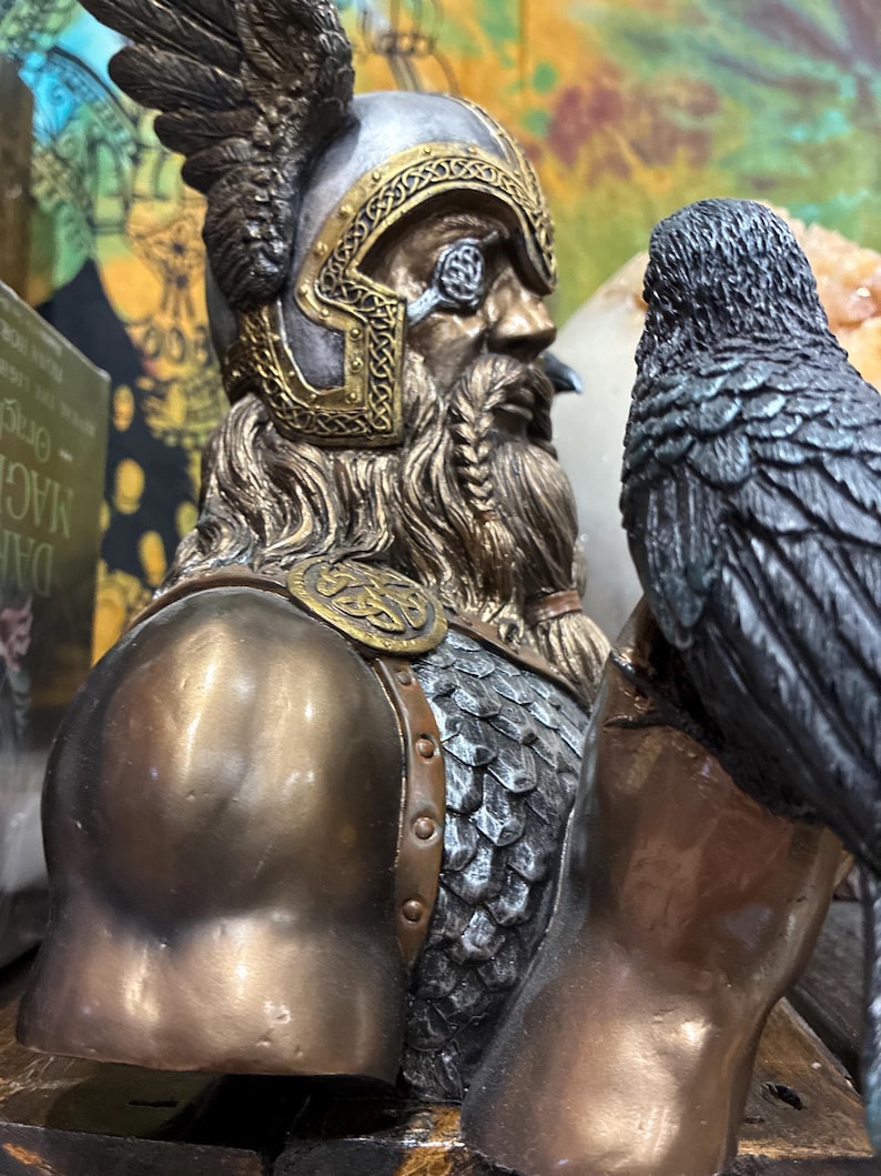 May include: A bronze-toned bust of a Viking warrior with a detailed helmet and beard. A raven sits on the warrior's shoulder. The bust has intricate gold-colored accents and a weathered appearance, suggesting a historical or fantasy theme.