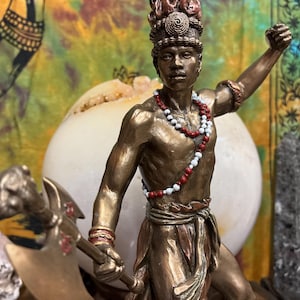 May include: Bronze-toned statue of a figure with a headdress and a necklace of red and white beads. The figure holds a weapon and raises one arm. The statue has intricate details and is set against a colorful background.