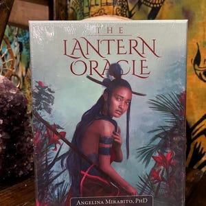 Puede incluir: Baraja de cartas The Lantern Oracle. La caja presenta una ilustración de una mujer con cabello largo y oscuro, adornada con un tocado, rodeada de flores rojas y follaje. El texto incluye "The Lantern Oracle" y los nombres del autor y el ilustrador.
