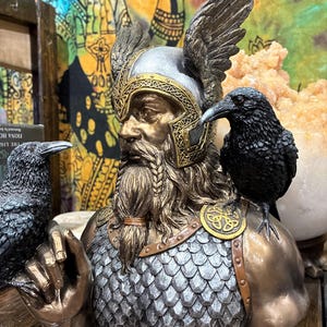 May include: A bronze-toned bust of a figure with a helmet and braided beard, adorned with two black raven statues. The figure wears a silver helmet with gold accents and has a chainmail chest. The bust is a decorative item.