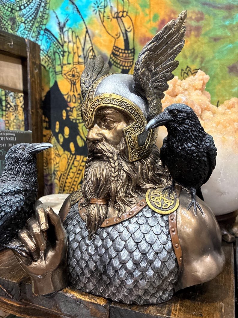 May include: A bronze-toned bust of a figure with a helmet and braided beard, adorned with two black raven statues. The figure wears a silver helmet with gold accents and has a chainmail chest. The bust is a decorative item.