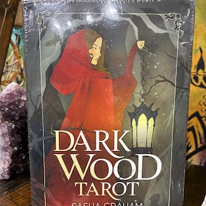 May include: A sealed tarot card deck titled "Dark Wood Tarot" featuring an illustration of a person in a red cloak holding a lantern. The deck's cover has the text "Dark Wood Tarot" and the names "Sasha Graham" and "Art by Abigail Larson".