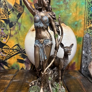 May include: Bronze statue of Artemis, the Greek goddess of the hunt, with a bow and arrow. The statue features a deer and is set on a base with the word "ARTEMIS". The statue is a detailed representation of the goddess.