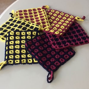 Puede incluir: Cinco agarraderas de olla de ganchillo en un esquema de color amarillo, negro y burdeos. Las agarraderas de olla están hechas con un patrón cuadrado y tienen un lazo para colgar.