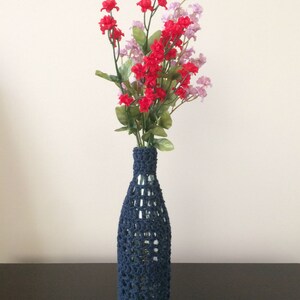 May include: A blue crocheted bottle cover with a glass bottle inside. The bottle is filled with a bouquet of red and pink flowers.