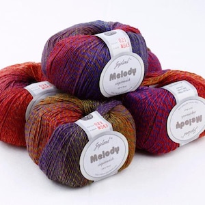 Peut inclure: Un groupe de cinq écheveaux de laine Jojoland Melody superwash. Chaque écheveau présente une palette de couleurs variées composée de violet profond, d'orange vif et de jaune doré. Des étiquettes en papier blanc avec du texte entourent chaque écheveau.