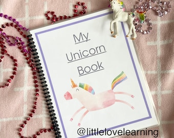 May include: A pink and white unicorn book with a rainbow mane and tail. The book has a purple spine and the text "My Unicorn Book" on the cover. The book is on a pink and white checked blanket with a small white unicorn figurine on top of the book.