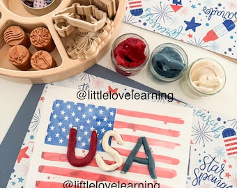 May include: A wooden craft tray filled with stamps, cookie cutters, and small jars of red, blue, and cream-coloured playdough. A watercolour painting of the American flag with the word "USA" made of playdough is also present.