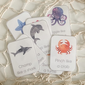 Op de afbeelding: Een verzameling educatieve flashcards met aquarel illustraties van zeedieren. Elke kaart toont een dier, zoals een haai of octopus, met een actie-prompt zoals "Chomp like a shark". De kaarten zijn wit met afgeronde hoeken.