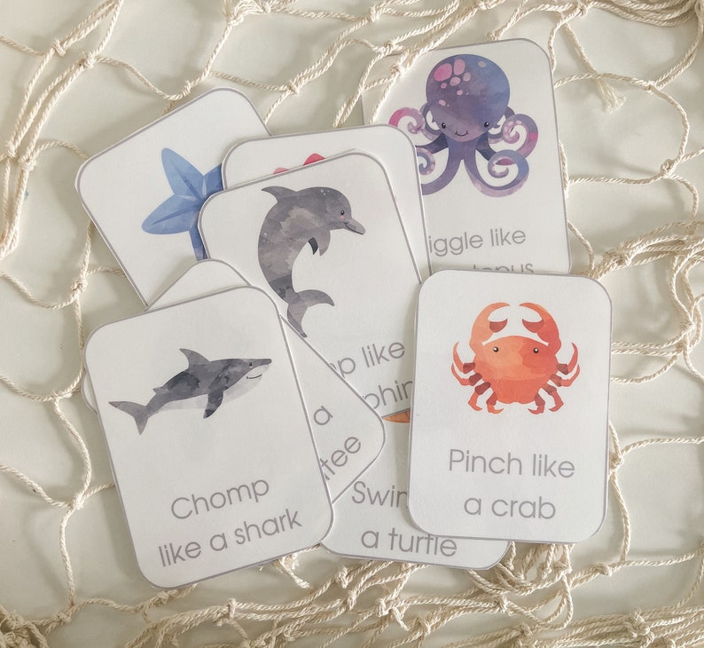 May include: A set of flashcards with watercolour illustrations of sea creatures. Each card shows an animal, like a shark or crab, and an action prompt, such as "Chomp like a shark" or "Pinch like a crab". The cards are white with rounded corners.