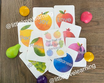 May include: A set of colourful cards with fruit illustrations and words representing the fruits of the spirit. The words on the cards include: Joy, Love, Peace, Patience, Kindness, Goodness, Faithfulness, Gentleness, and Self-Control. The cards are on a wooden surface with small colourful play dough balls.